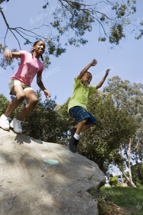 Preview: Boy (6-8) And Girl (12-14) Leaping From Rock In Park, Smiling, Low Angle View