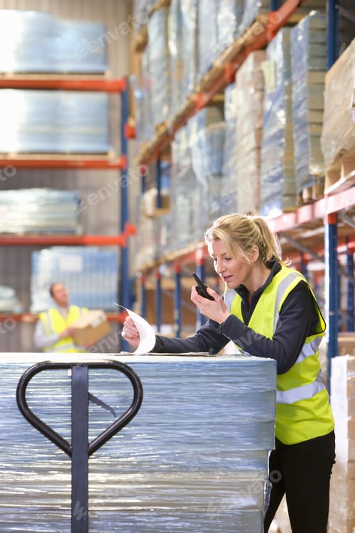Preview: Female Warehouse Worker Using Walkie-Talkie And Viewing Paperwork On Pallet Truck