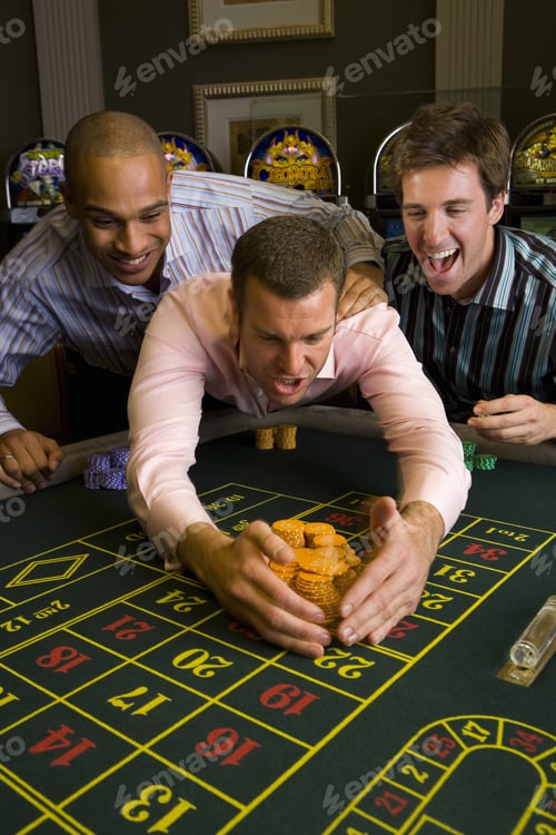 Preview: Young Man With Friends Collecting Pile Of Gambling Chips From Roulette Table In Casino, Smiling