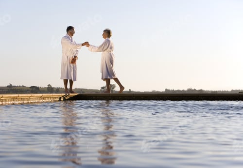 Preview: Senior Couple Wearing White Bath Robes, Standing Outdoors By Swimming Pool, Side View