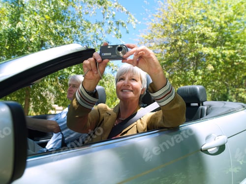 Preview: Senior Couple Driving Convertible Car On Summer Road Trip In Countryside And Taking A Picture