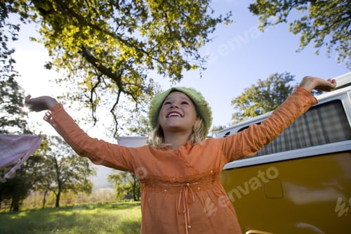 Preview: Girl With Arms Raised By Camper Van In Field, Smiling, Low Angle View