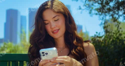 Preview: Smiling Brunette Typing Message On Phone Sitting On Park Bench Closeup. Pretty Asian Woman Chatting