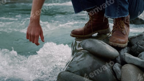 Preview: Closeup Man In Hiking Boots Sitting On Rock At Mountain Stream. Male Hand Touching Fresh Water From