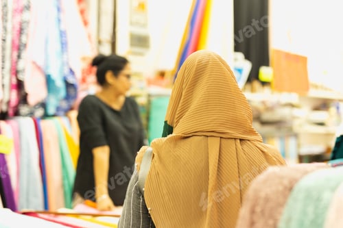 Preview: Back View Of Muslim Woman With Cream Hijab Choosing Fabrics At Market.