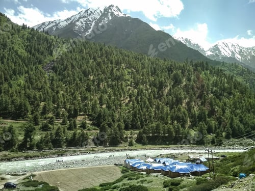 Preview: A Beautiful View Of Baspa River Surrounded By Green Coniferous Trees And Snow Capped Mountains