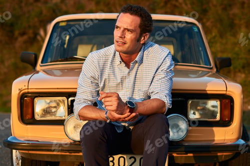 Preview: A Closeup Shot Of A Man Sitting In Front Of His Old Vintage Car. Serious Look On A Curly Model Face