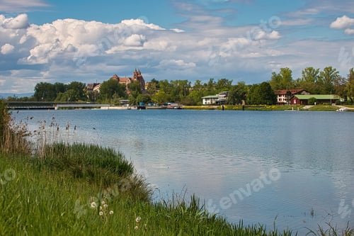 Preview: A Landscape Of A Lake Surrounded By Greenery And Buildings Under The Sunlight