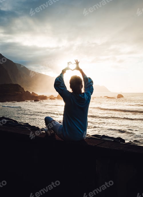 Preview: A Silhouette Of A Girl Sitting By The Sea Making A Heart With Her Fingers Against An Orange Sunset