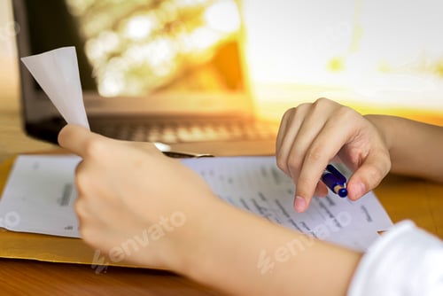 Preview: Business Woman Hand With Pen And Reading Piece Of Paper Documents With Laptop In Background
