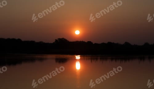 Preview: A Closeup Shot Of A Lake With Silhouettes Of Trees And The Round Sun In The Sky At Sunset