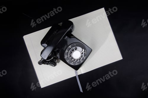 Preview: Overhead View Of A Black Rotary Telephone Resting On A Notebook On A Black Background