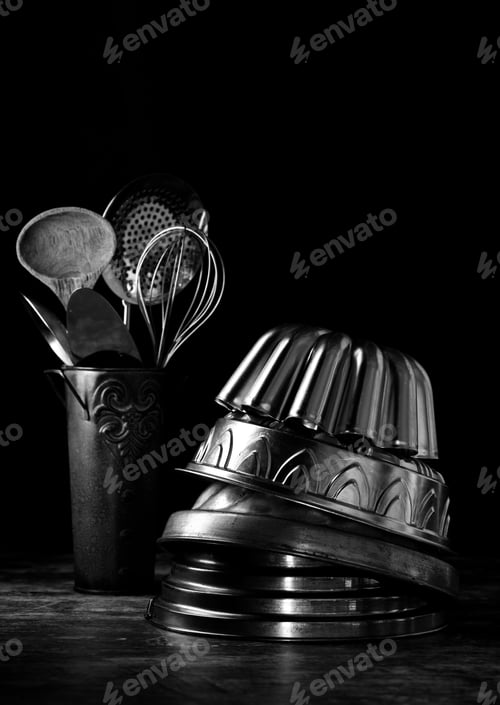 Preview: Black And White Vertical View Of A Stack Of Vintage German Bundt Cake Pans And A Tin Of Utensils In