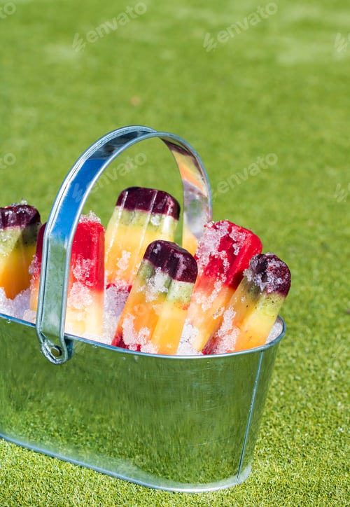 Preview: A Close Up Of A Tin Pail Filled With Snow And Rainbow Popsicles Against A Green Grass Background