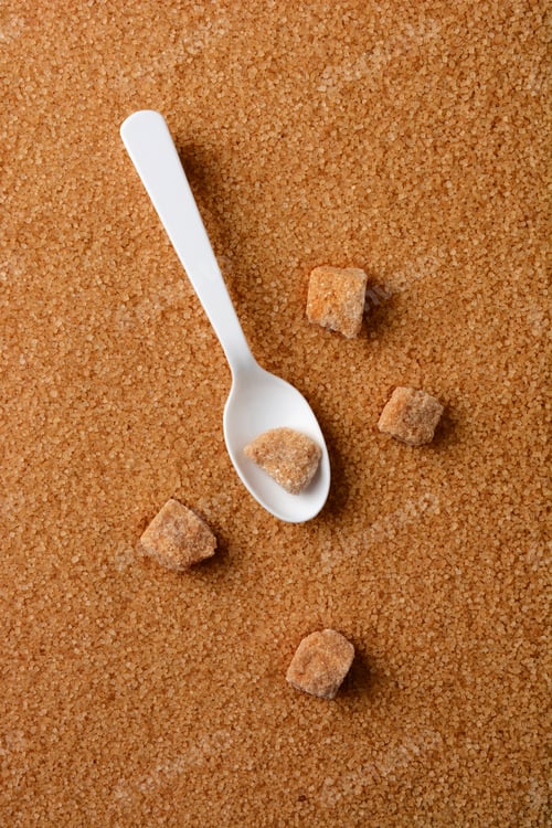 Preview: Overhead Shot Of Lumps Of Raw Brown Sugar And A White Spoon Laying Of A Bed Of Granulated Turbinado