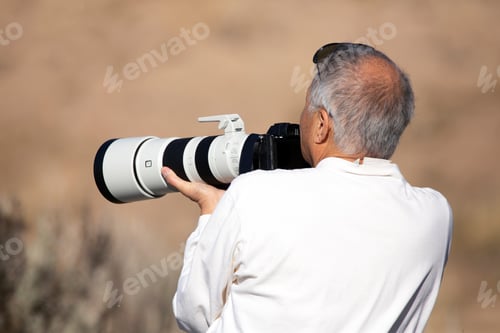 Preview: Older Man Holding A Large Camera With A White Telephoto Lens In The Desert