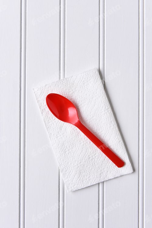 Preview: Top View Of A Red Plastic Spoon On A White Napkin On A White Table. Vertical Format With Copy Space.
