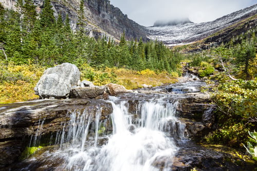 Vorschau: Wunderschöner Wasserfall, der sich über Felsen mit goldenen Herbstblättern im Hintergrund im Glacier Nap erhebt