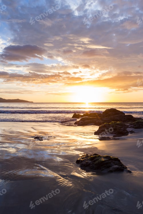 Preview: Colorful Sunset Reflecting On The Waves And Wet Sand With A Grouping Of Rocks In The Bottom Of The