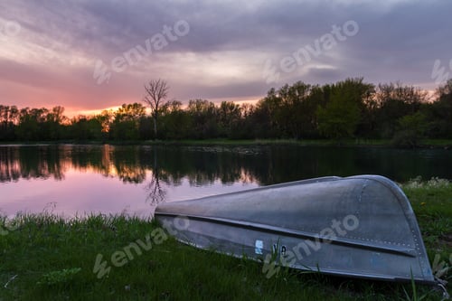 Preview: Sunset Reflecting On A Pond With A Small Paddle Boat On The Green Grassy, Shore