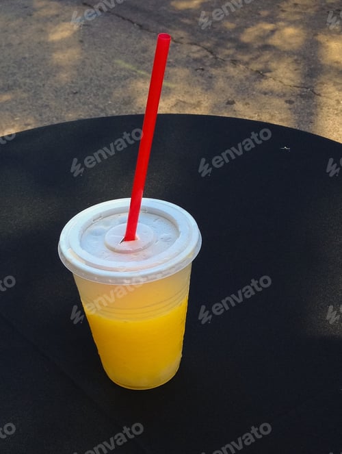 Preview: Yellow Drink In A Transparent Plastic Cup With Cover And A Red Straw On A Black Round Table