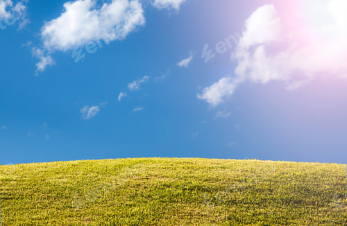 Preview: Bright Green Sunlit Grass Hill With Blue Sky And Clouds With Sun Flare And Copy Space