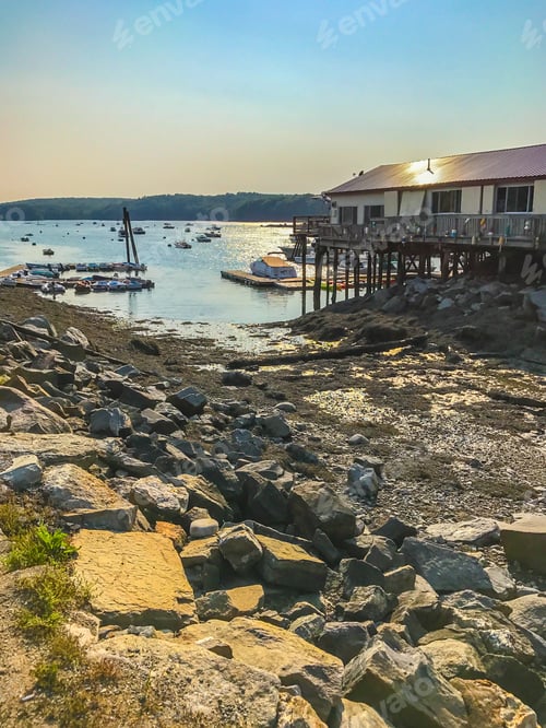 Preview: A Building On Pilings Over The Water Of A Bay With The Rocky Shore In The Foreground And Boats And