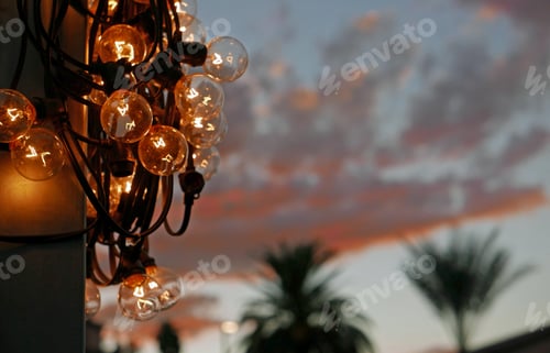 Preview: String Of Glowing Light Bulbs Wrapped Around A Pole In Front Of An Evening Sky With Red Clouds