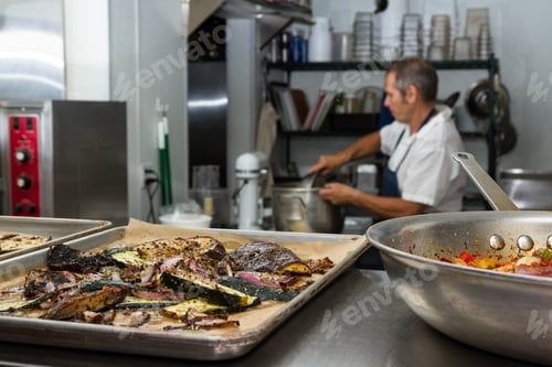 Preview: Roasted Vegetables On Trays With A Chef Preparing For Diner Service In The Background