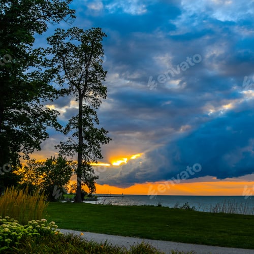 Preview: The Sun Setting Over Lake Erie With Trees And The Shoreline In The Foreground And Thick Clouds In