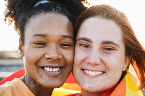 Preview: Multiracial Women Gay Couple Smiling On Camera Wearing Rainbow Flag Outdoor - Focus On Faces