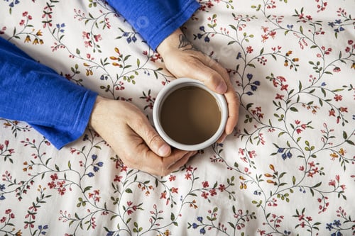 Preview: A Closeup Top View Of A Tattooed Man'S Hands Holding A Cup Of Coffee In Bed On A Floral Blanket