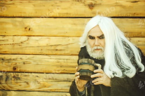 Preview: Druid Old Man With Long Grey Hair And Beard On Serious Face With Wooden Mug In Hands Outdoor On