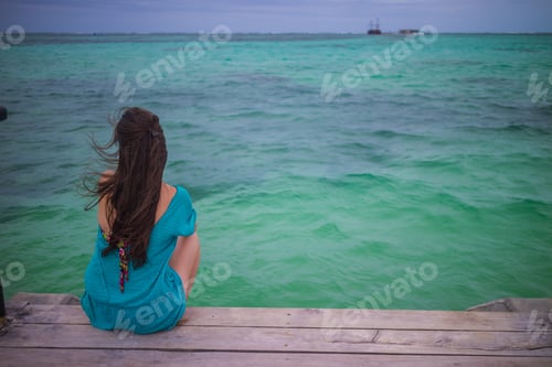 Preview: Closeup Back View Of Long Hair Woman Sitting In Blue Beach Dress Looking Out Towards Blue Ocean And