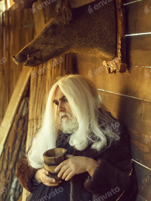 Preview: Old Druid Man With Long Silver Hair And Beard Holds Wooden Mug On Stuffed Boar Head Background