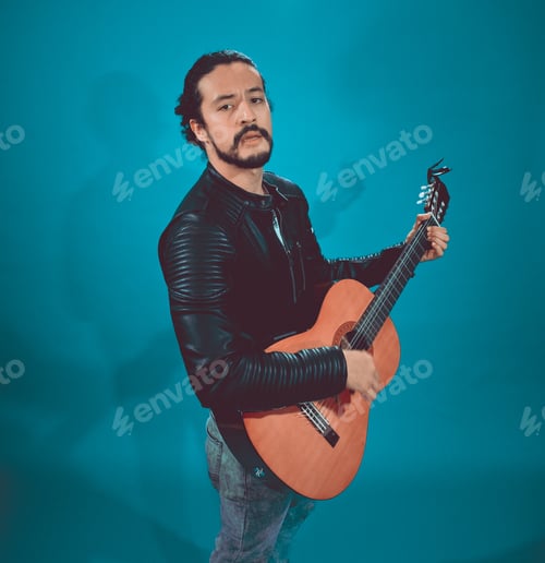 Preview: Portrait Of Attractive Latin Young In Rocker Jacket, On Blue Background,With Acoustic Guitar