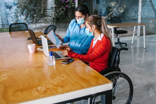 Preview: Young Latin Woman In Wheelchair Working With Another Woman Colleague At Workplace Or Office In