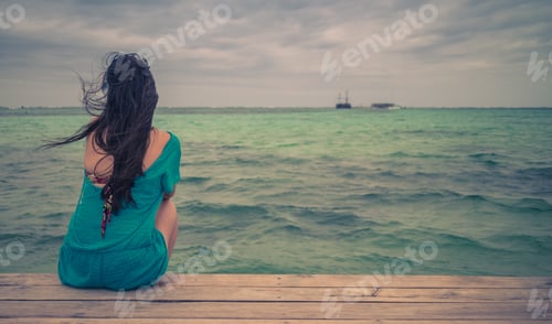 Preview: Closeup Back View Of Long Hair Woman Sitting In Blue Beach Dress Looking Out Towards Blue Ocean And