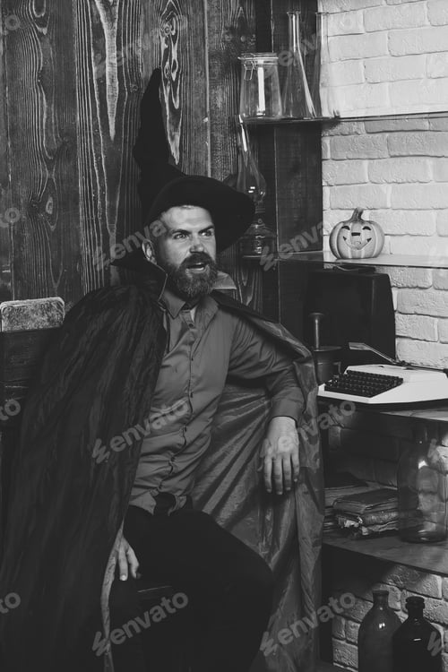 Preview: Halloween Man In Witch Hat And Cloak. Wizard Sitting At Pumpkin, Typewriter, Books On Shelves