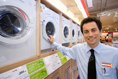 Preview: Young Salesman Wearing A Name Tag And Standing By A Row Of New Washing Machines And Ready To Help