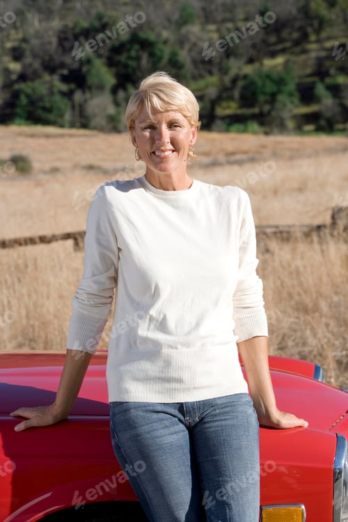 Preview: Vertical Three Quarter Portrait Of A Joyous Senior Woman Leaning Against A Red Car On A Sunny Day.