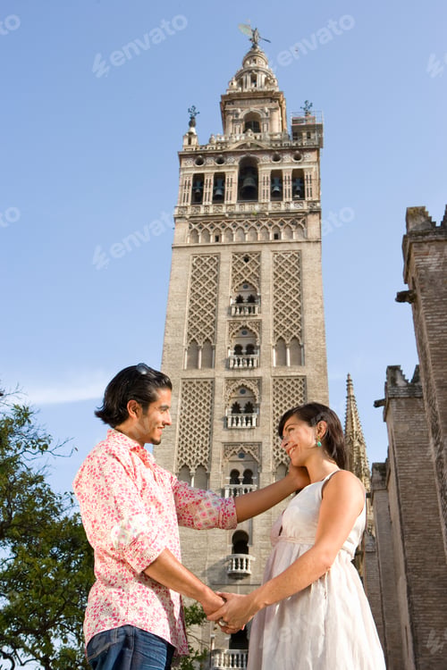 Preview: Low Angle View Of A Tourist Couple Holding Hands On A Sunny Day With A Tower In The Background.