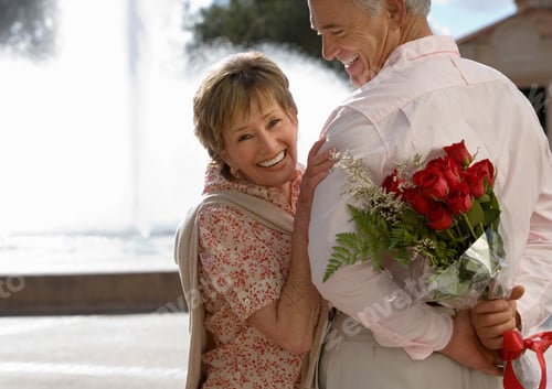 Preview: Close-Up Of A Senior Couple Standing Near A Water Fountain With The Man Holding Bouquet Of Red