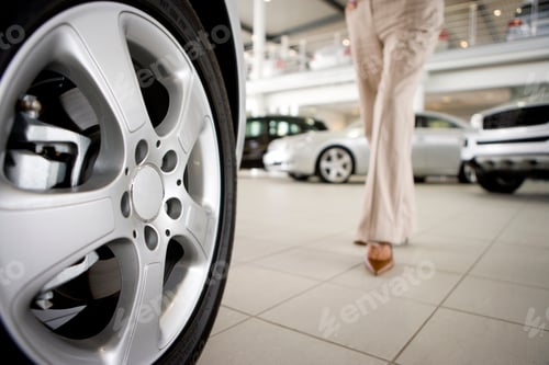 Preview: Close-Up Shot Of A Young Woman In Trousers Walking Towards A Car In The Showroom.