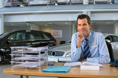 Preview: Portrait Of A Smiling Car Salesman Sitting At The Desk In A Car Showroom.