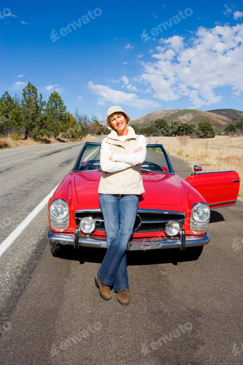 Preview: Vertical Portrait Of A Mature Woman Standing In Front Of A Red Convertible On A Country Road Smiles