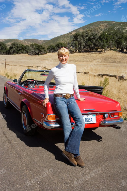 Preview: Vertical Portrait Of A Mature Woman Leaning Over A Red Convertible On A Country Road Smiles At The