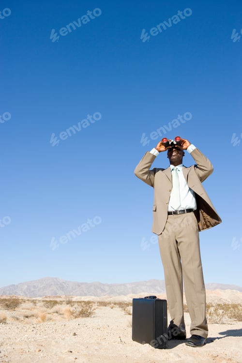 Preview: Low Angle View Of A Businessman Standing By A Briefcase Using Binoculars In The Desert.