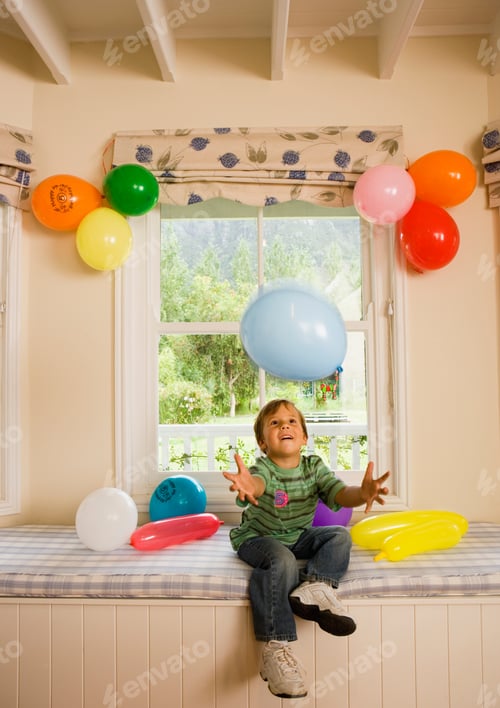Preview: Vertical Shot Of A Young Boy Sitting On A Window Seat At Home And Catching A Falling Party Balloon.