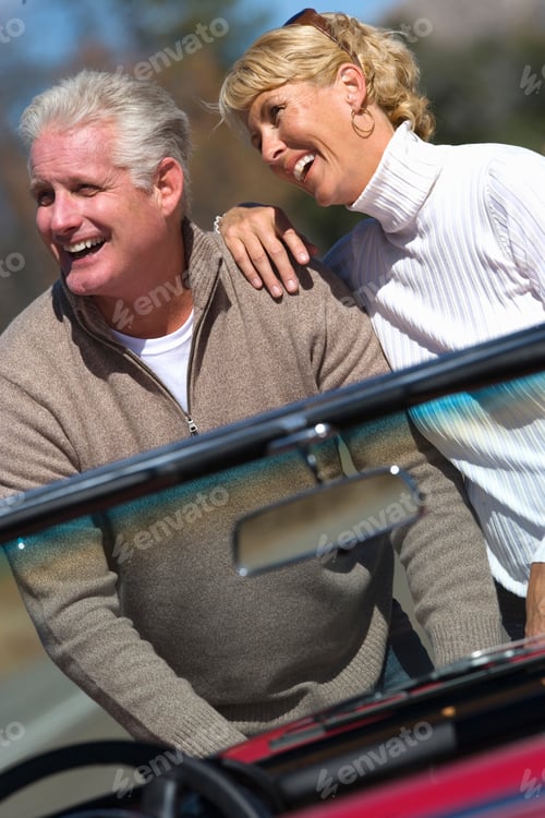 Preview: Vertical Tilted Waist Up Portrait Of A Joyous Senior Couple In Front Of Bonnet Of Their Red
